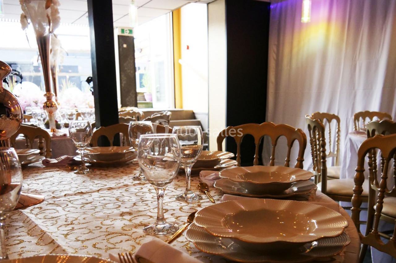 Table dressée avec vaisselle élégante, nappes dorées et chaises en bois dans une salle de réception lumineuse.