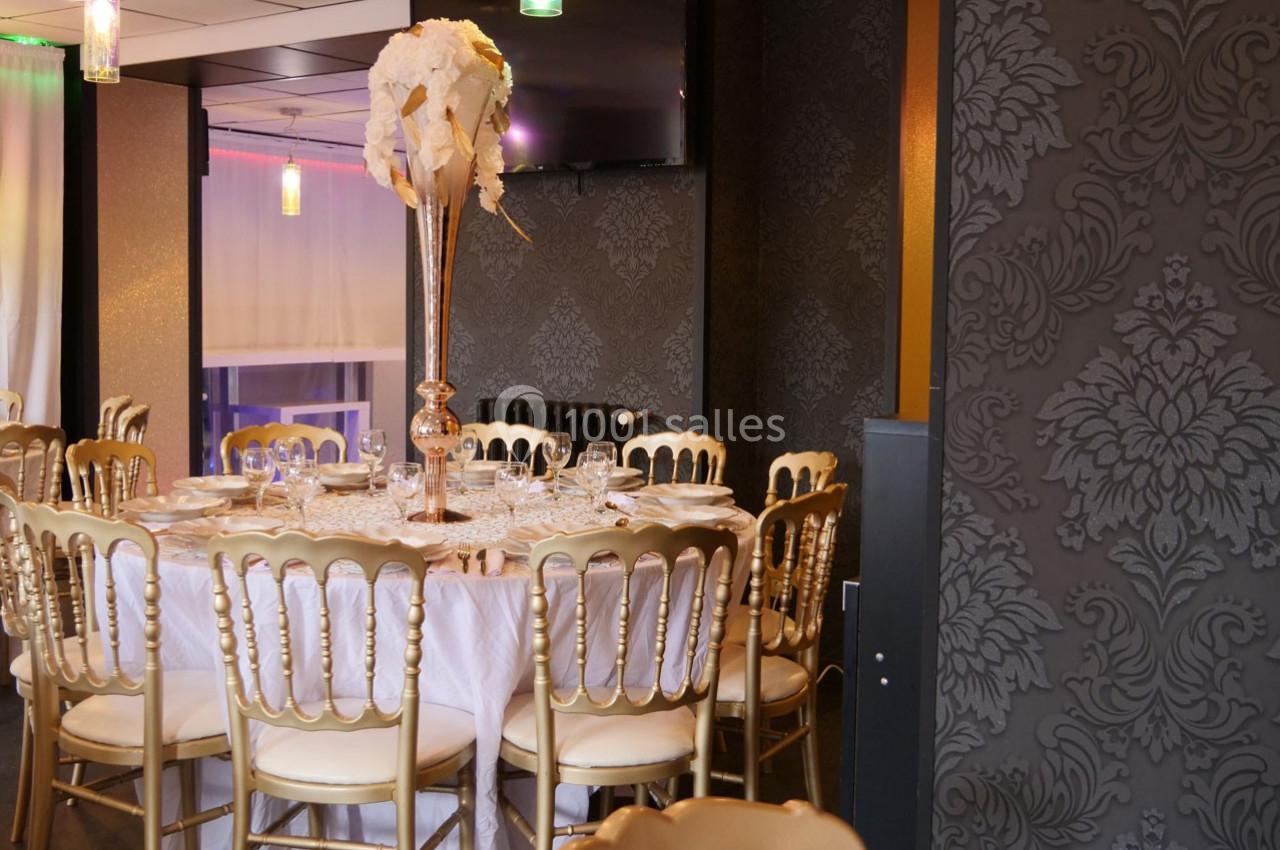 Salle de réception élégante avec table ronde dressée, chaises dorées et décoration florale sur un vase haut.