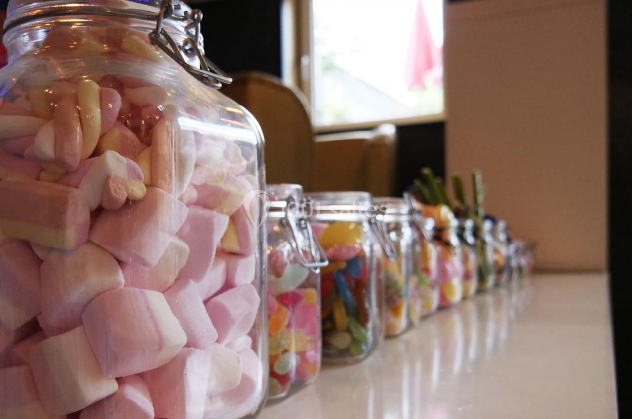 Bocaux en verre alignés sur une table, remplis de bonbons colorés, dont des guimauves roses au premier plan.
