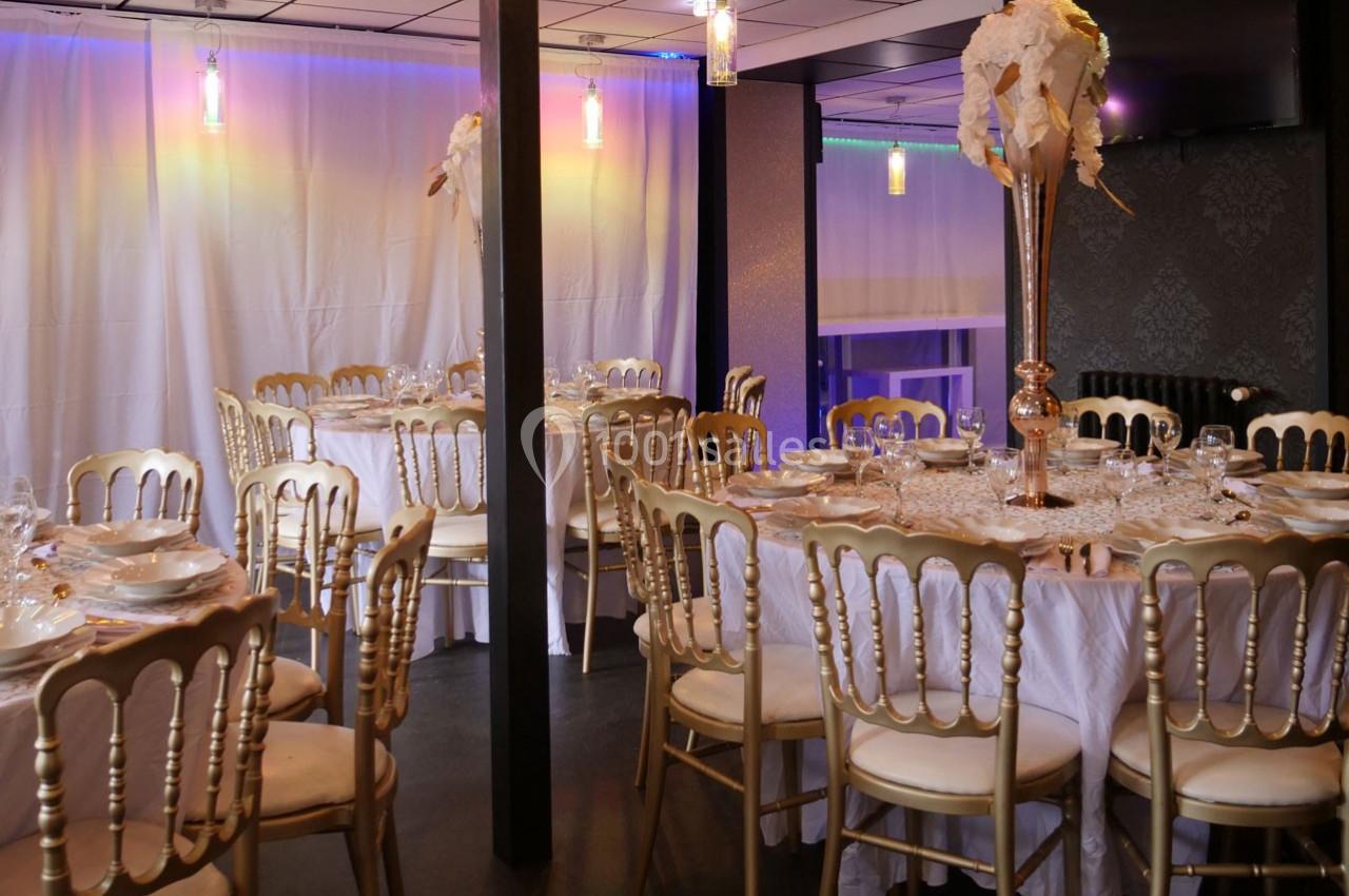 Salle de réception décorée avec des tables rondes, nappes blanches, chaises dorées et centre de table floral.