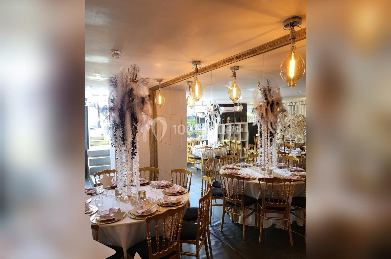 Salle de réception décorée avec des tables rondes, nappes blanches, chaises dorées et centres de table ornés de plumes.