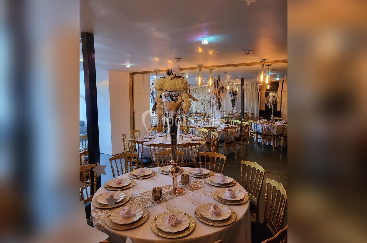 Salle de réception décorée avec des tables rondes élégantes, nappes blanches, chaises dorées et centre de table floral.