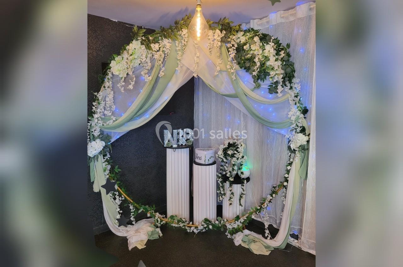 Arche circulaire décorée de fleurs blanches et feuillage, avec rideaux lumineux en arrière-plan et colonnes blanches.