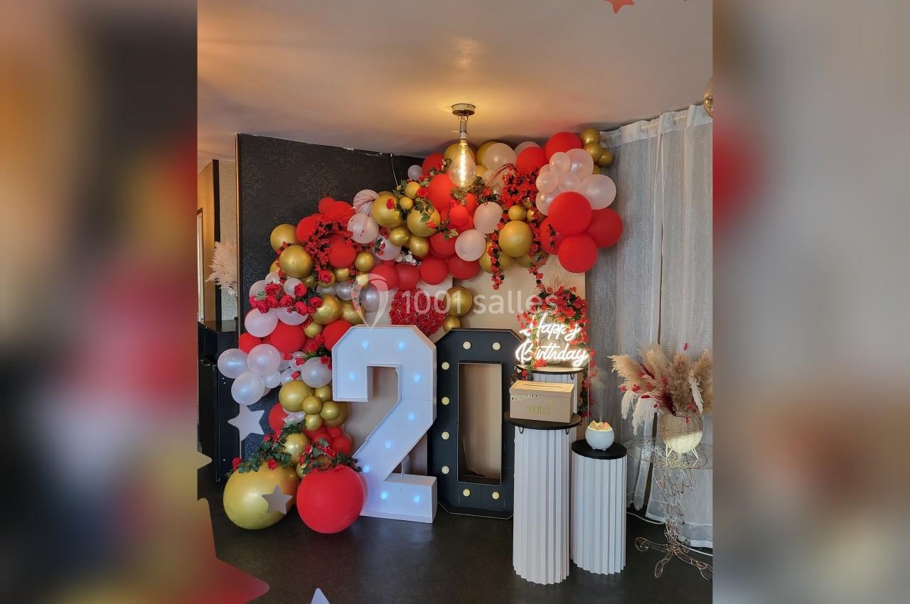 Décoration festive avec ballons rouges, blancs et dorés, chiffres lumineux ’20’ et petit gâteau sur des colonnes.