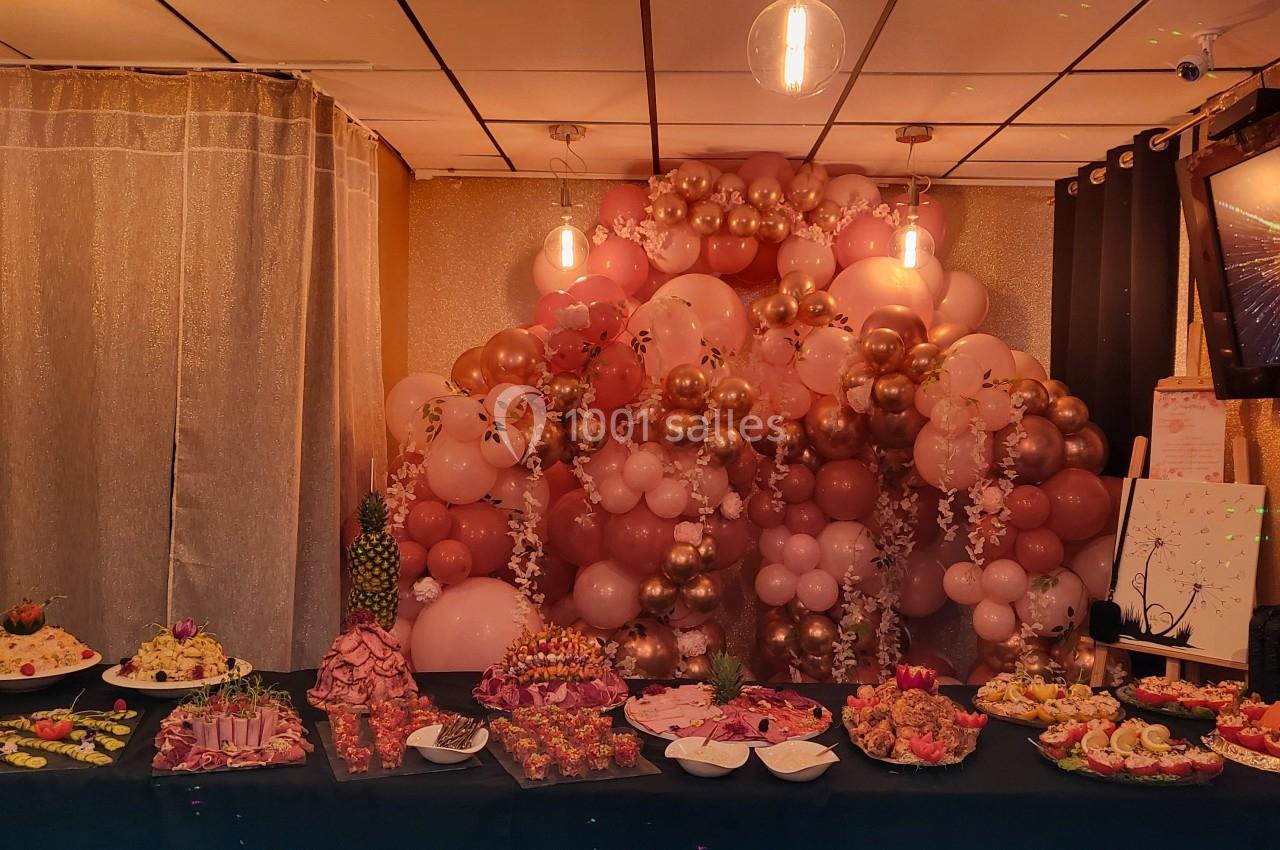 Table garnie de plats variés devant une décoration murale composée de ballons roses et dorés sous un éclairage tamisé.
