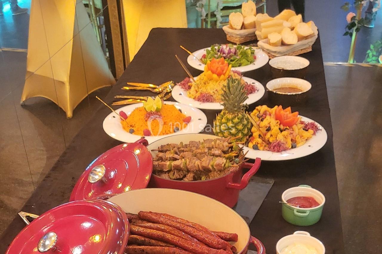 Buffet avec plats variés : brochettes, saucisses, salades colorées, pain et sauces, présenté sur une table noire.