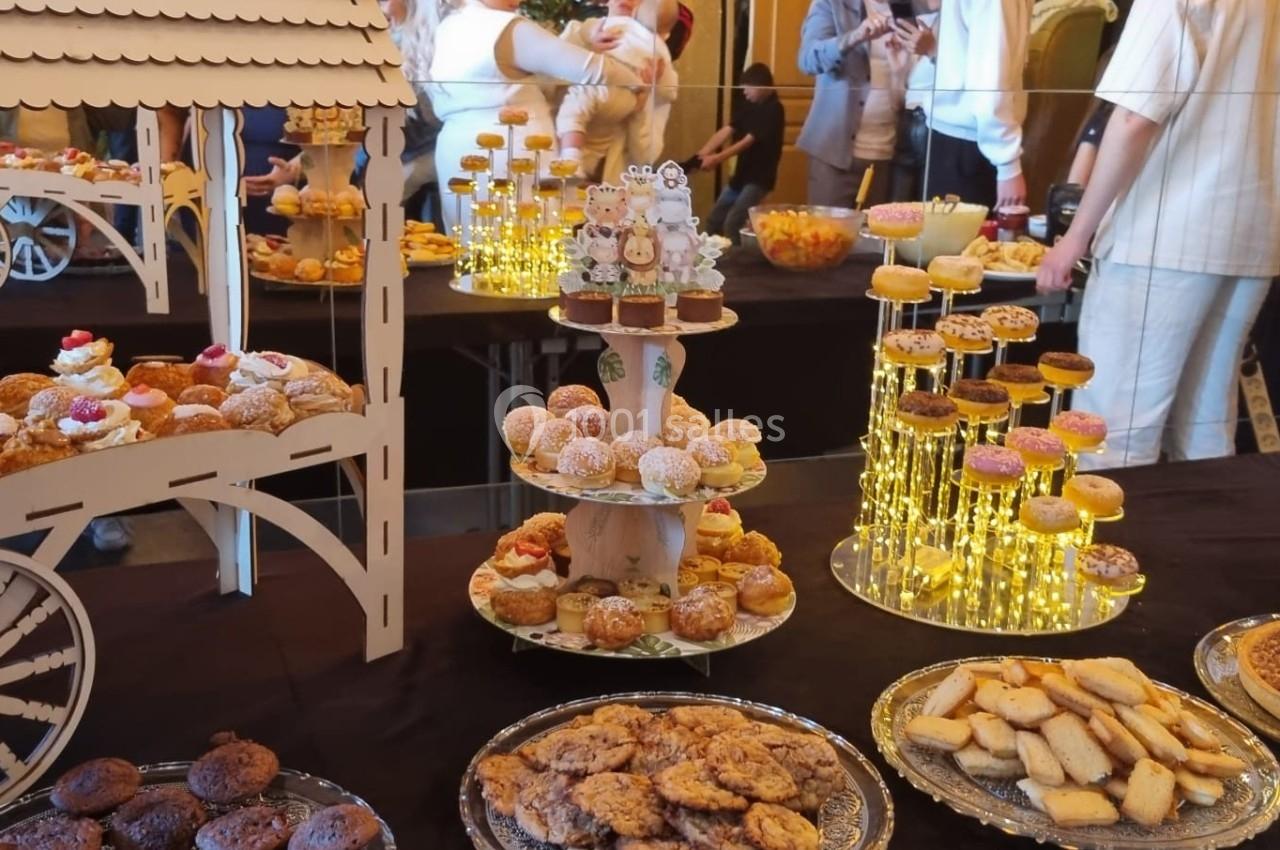 Table garnie de pâtisseries variées, présentées sur des plateaux et supports décoratifs, avec des invités en arrière-plan.