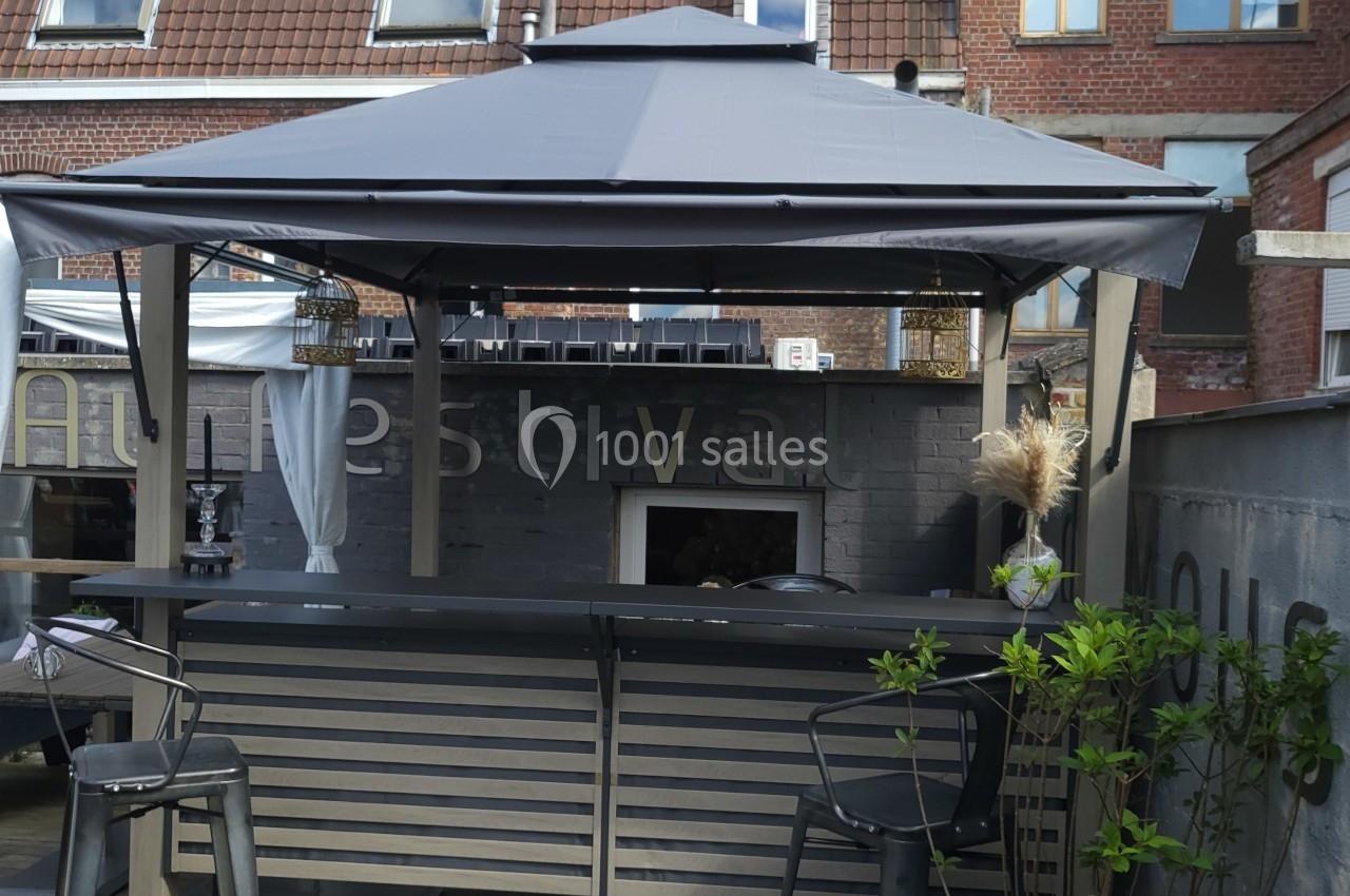 Bar extérieur avec un toit en toile grise, tabourets hauts et décorations végétales, devant un mur en briques.