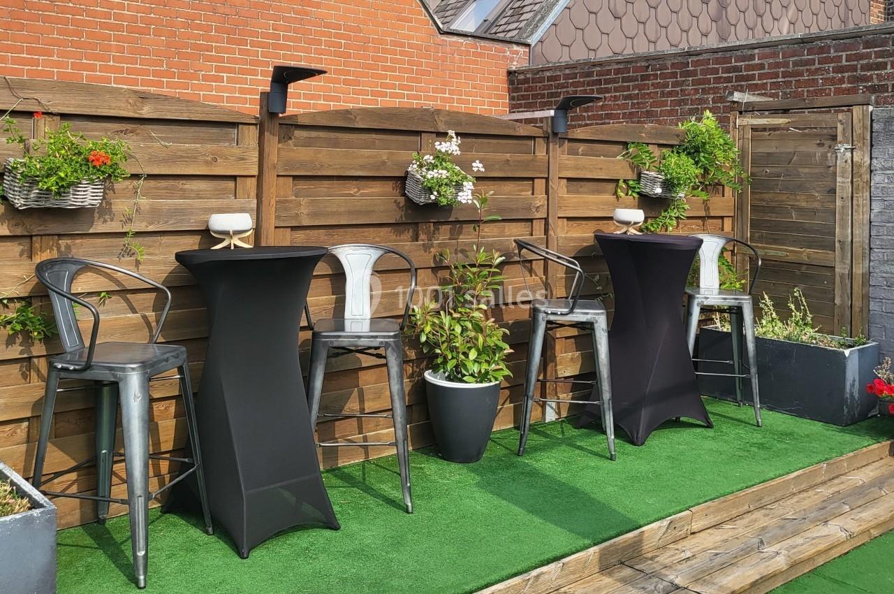 Tables hautes noires entourées de chaises métalliques sur une terrasse avec pelouse artificielle et décorations florales.