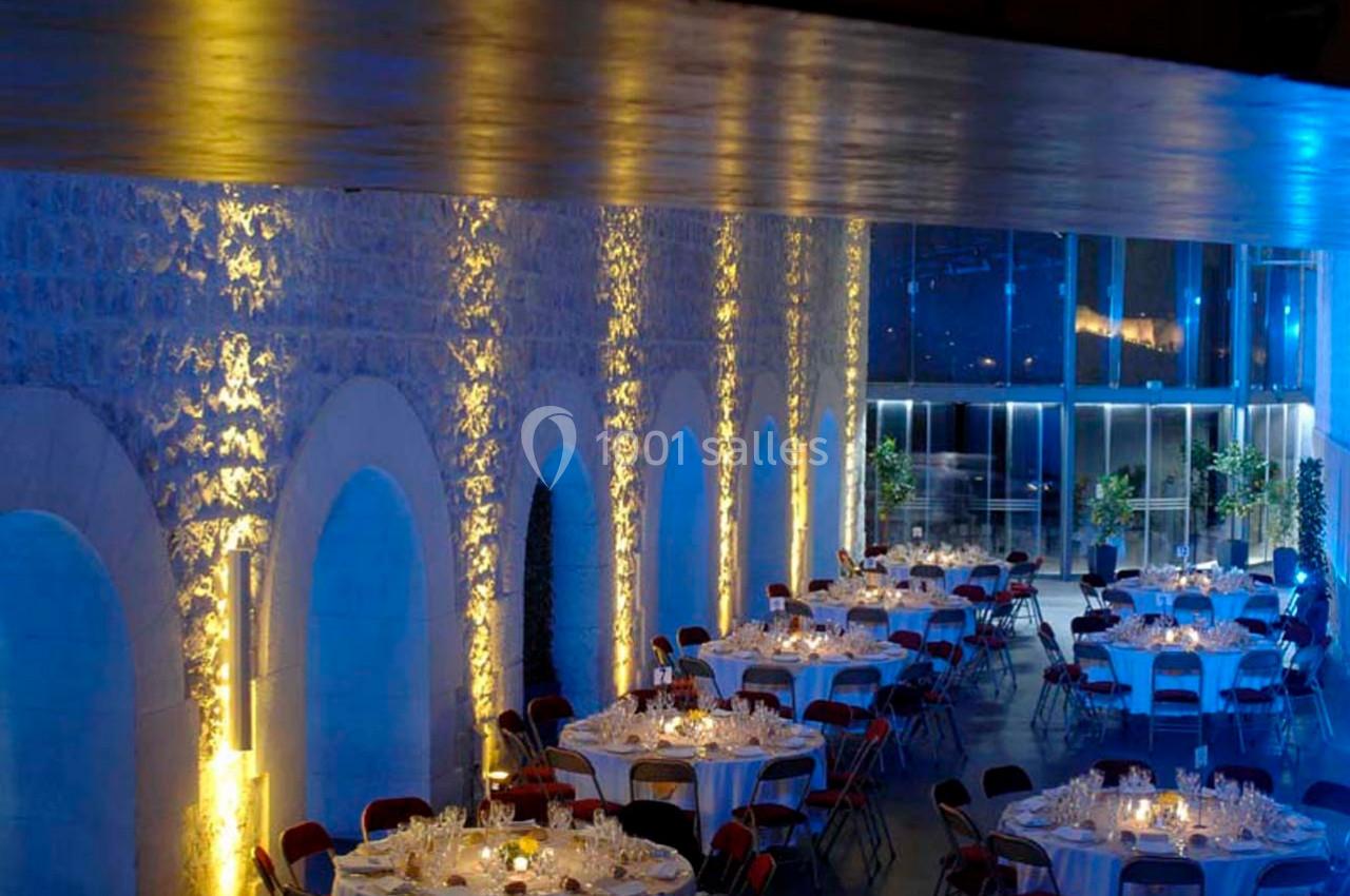 Salle de réception avec murs en pierre éclairés, tables rondes dressées et grandes baies vitrées en arrière-plan.