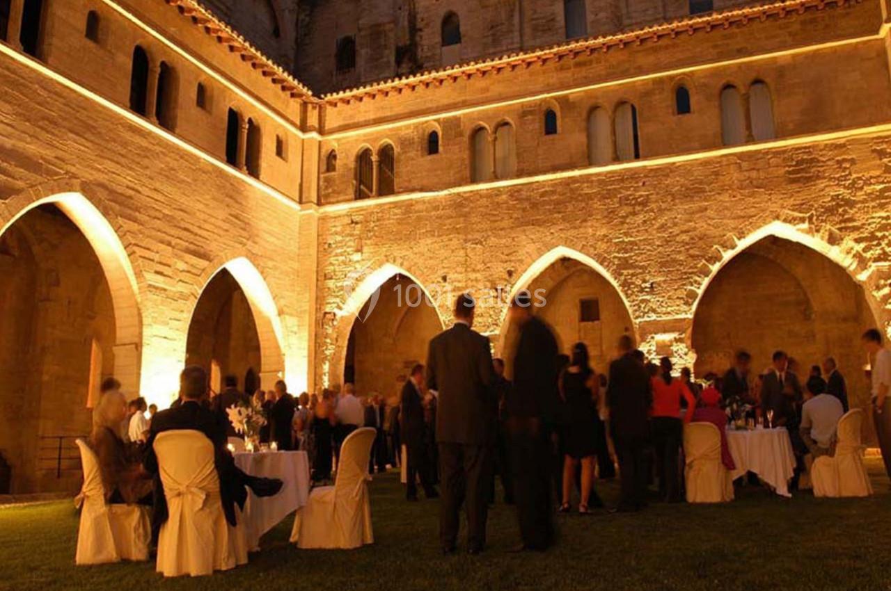 Personnes rassemblées dans une cour médiévale éclairée, assises ou debout autour de tables lors d'un événement en soirée.