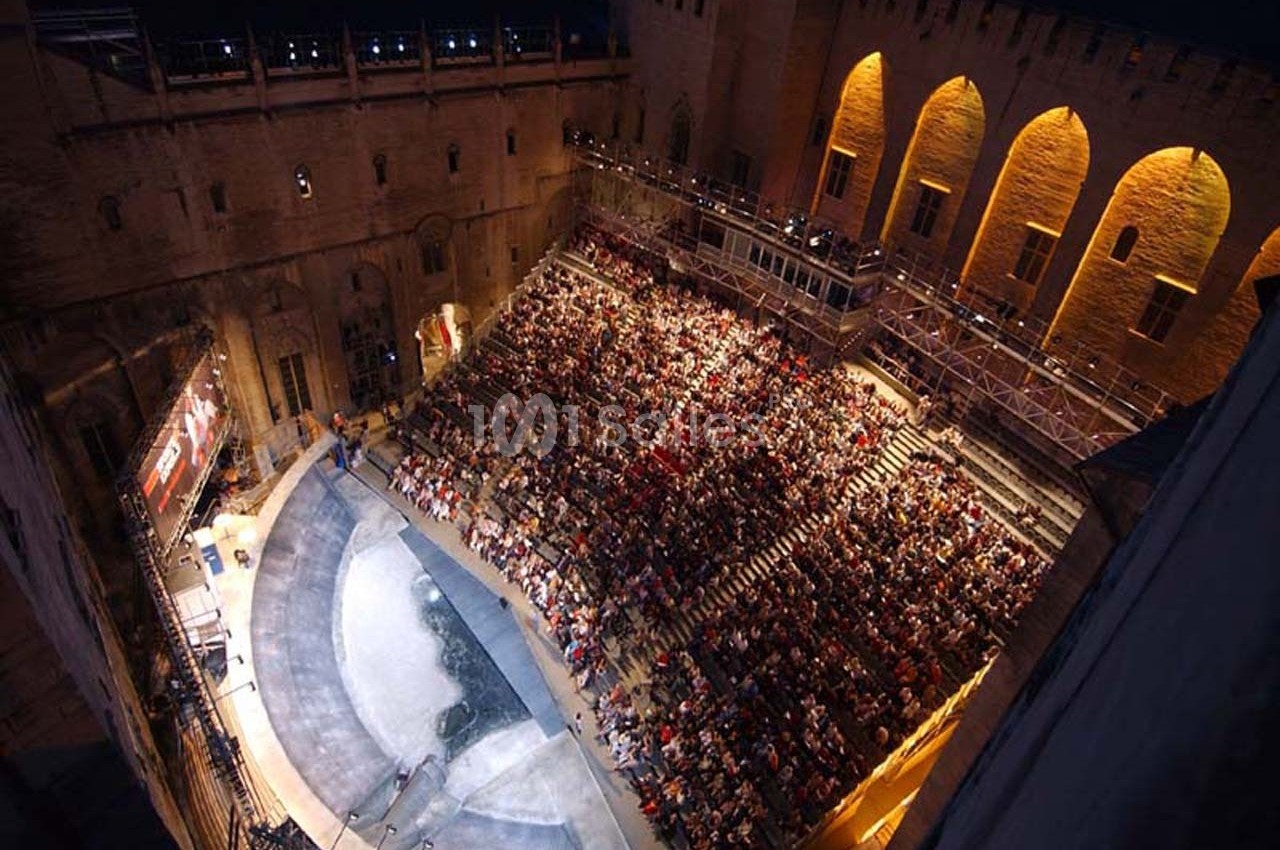 Vue d'une scène de théâtre en plein air, entourée de gradins remplis de spectateurs, dans un cadre architectural historique.