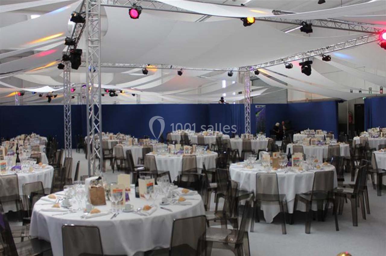 Salle de réception avec des tables rondes dressées pour un événement, sous une structure avec éclairage coloré.