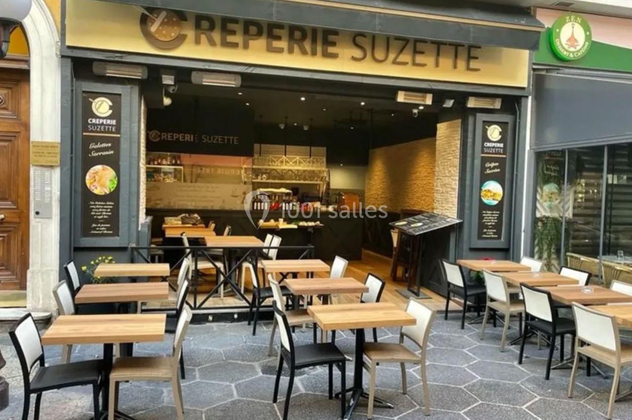Terrasse d'une crêperie avec tables et chaises en bois, devanture vitrée et enseigne ’Crêperie Suzette’.