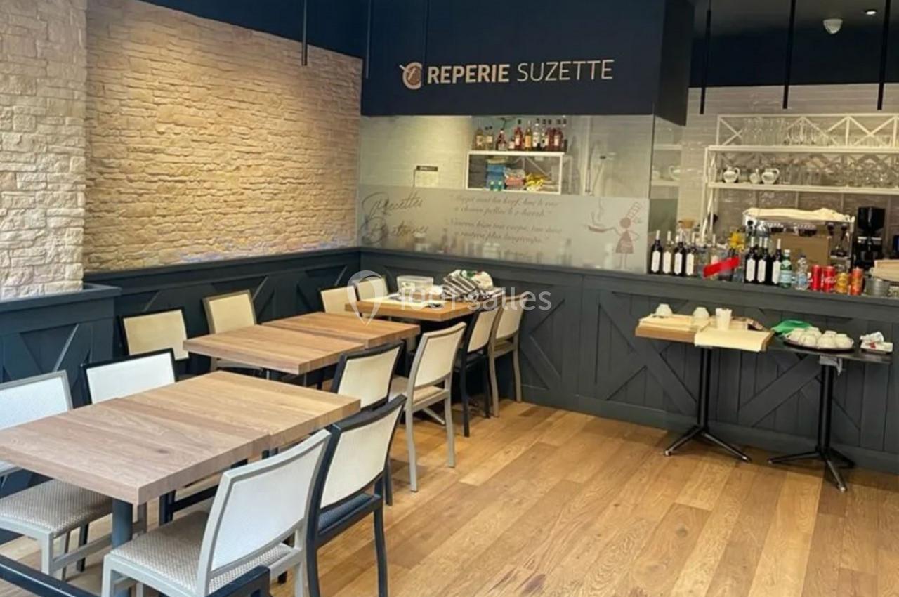 Salle de restaurant avec tables en bois, chaises claires, mur en briques et comptoir visible en arrière-plan.