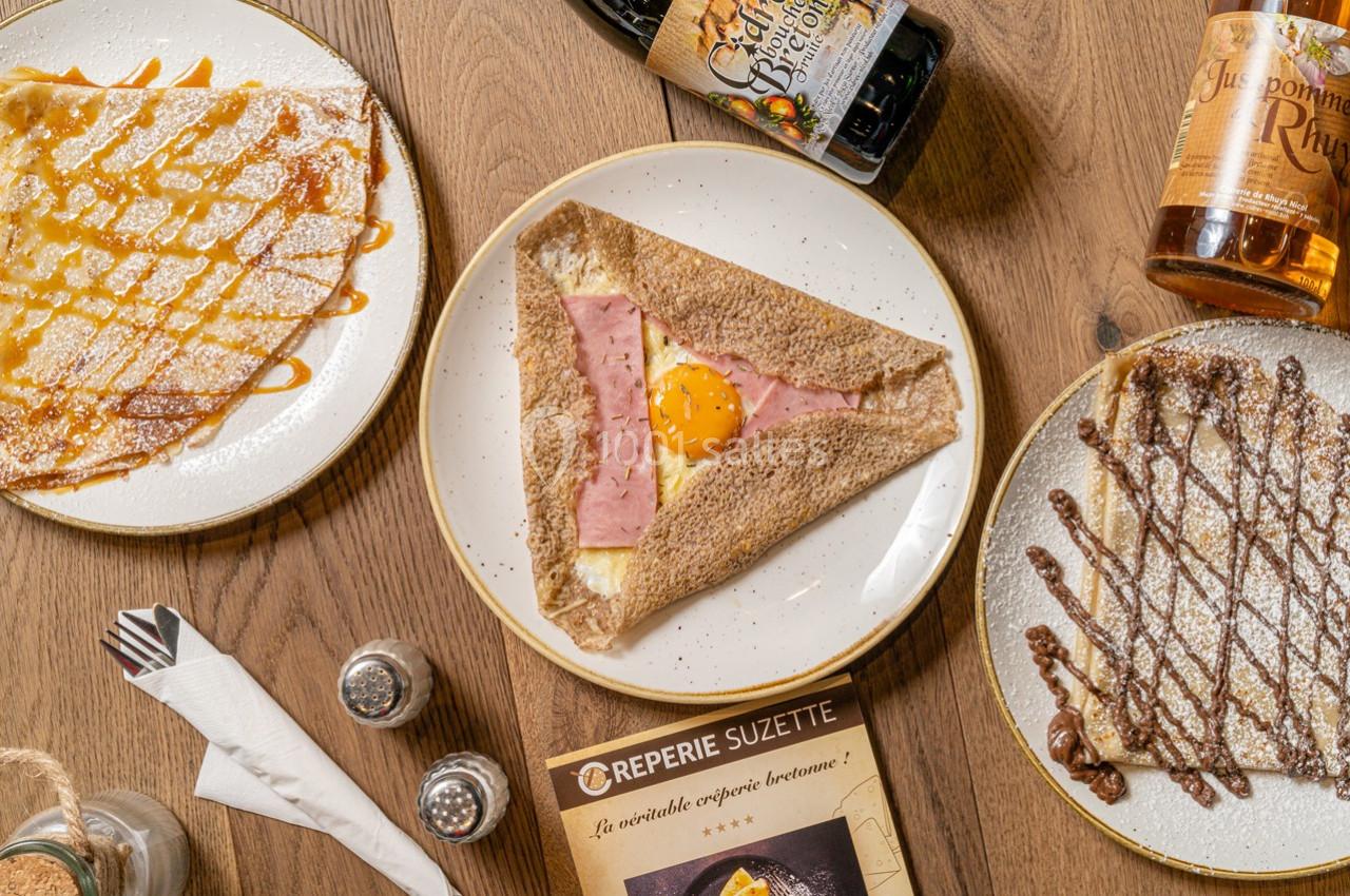 Assiette de galette au sarrasin garnie de jambon, œuf et fromage, entourée de crêpes sucrées et boissons sur une table en…