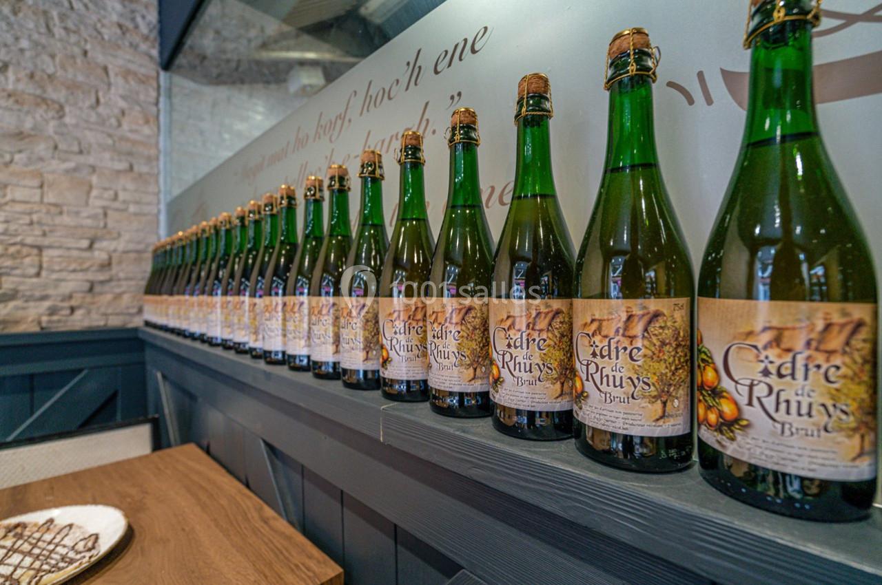 Alignement de bouteilles de cidre sur une étagère en bois, devant un mur en pierre et une vitre décorative.