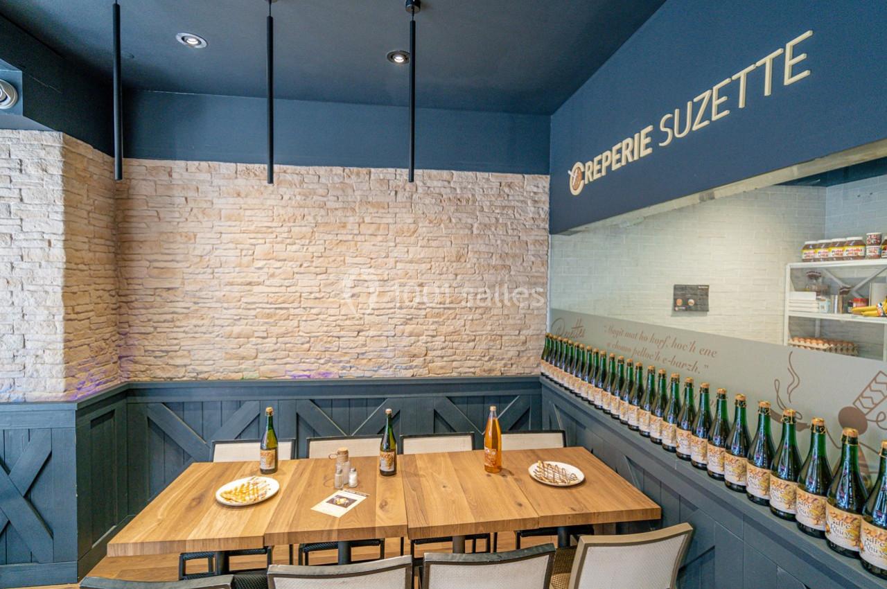 Salle de restaurant avec table en bois, chaises, bouteilles alignées sur une étagère et mur en pierre claire.