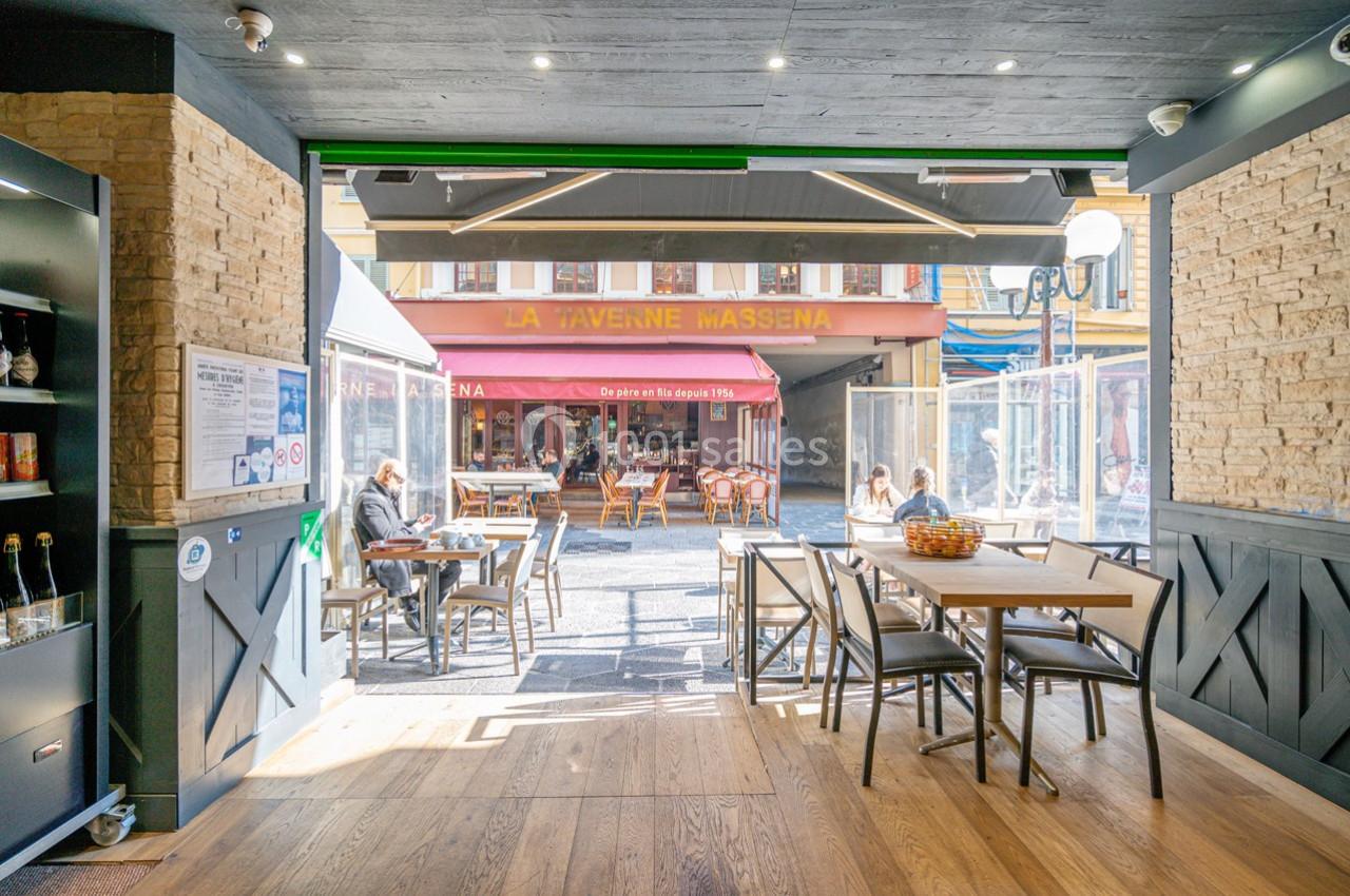 Vue depuis l'intérieur d'un café sur une terrasse ensoleillée et une rue animée avec des passants et des tables occupées.