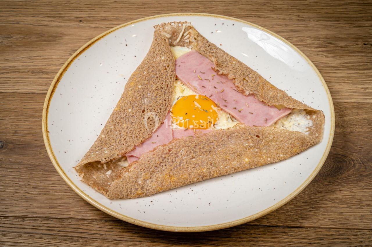 Crêpe de sarrasin garnie de jambon, fromage fondu et œuf au plat, servie sur une assiette en céramique.