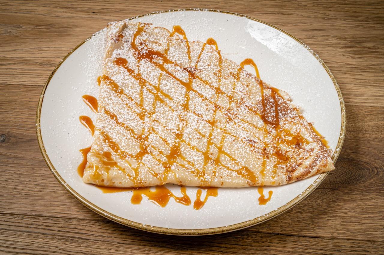 Crêpe pliée sur une assiette, garnie de caramel et saupoudrée de sucre glace, posée sur une table en bois.
