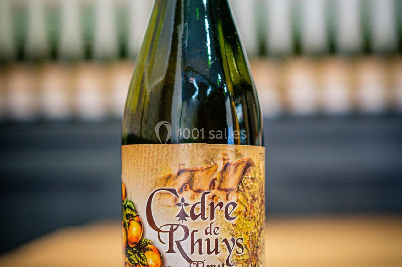 Bouteille de cidre brut avec une étiquette illustrée de pommes et un fond flou de bouteilles alignées.
