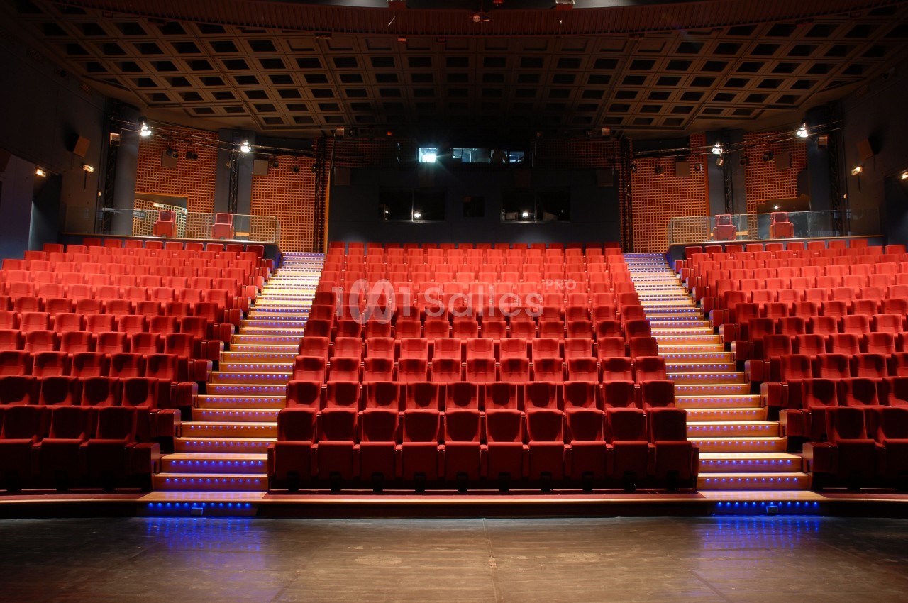 Salle de théâtre vide avec des rangées de sièges rouges et un éclairage tamisé.