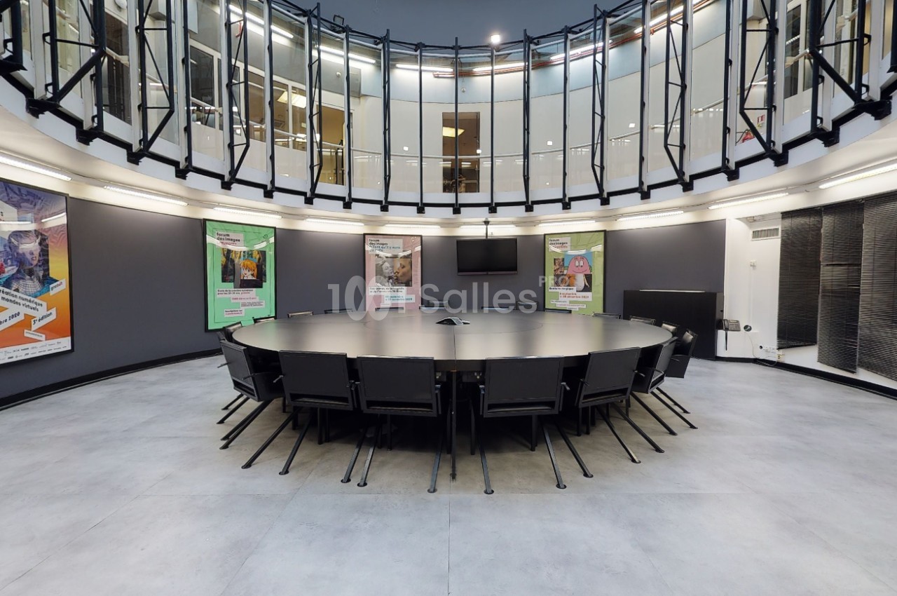 Salle de réunion circulaire avec une grande table noire entourée de chaises, éclairée par une verrière et des affiches…