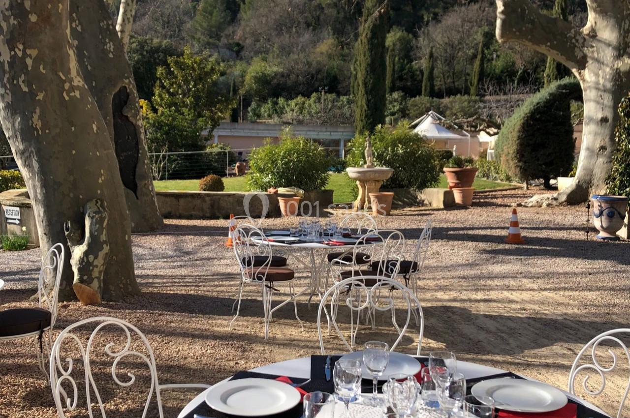 Terrasse extérieure avec tables dressées, chaises en fer forgé, arbres et végétation en arrière-plan.