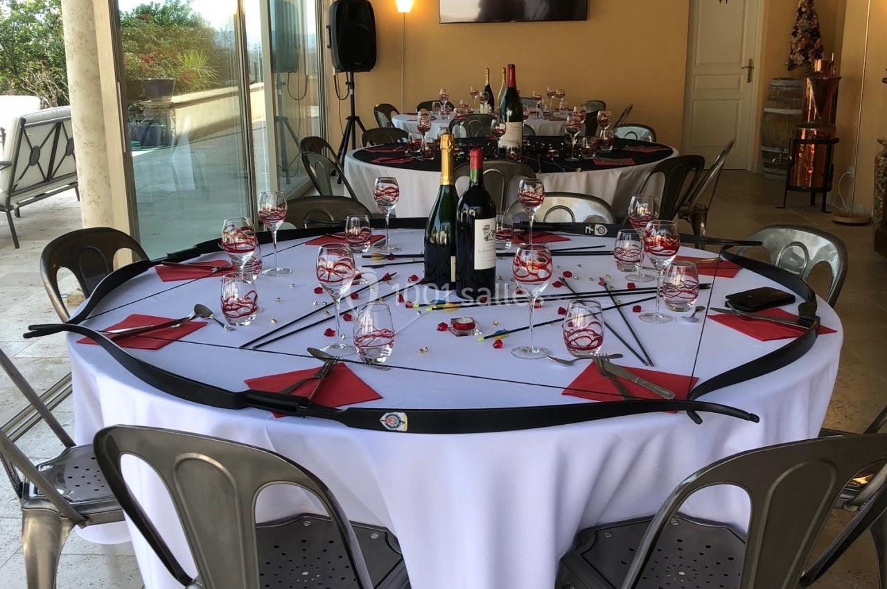 Table ronde dressée avec nappes blanches, verres, serviettes rouges et bouteilles, dans une salle lumineuse avec baies…