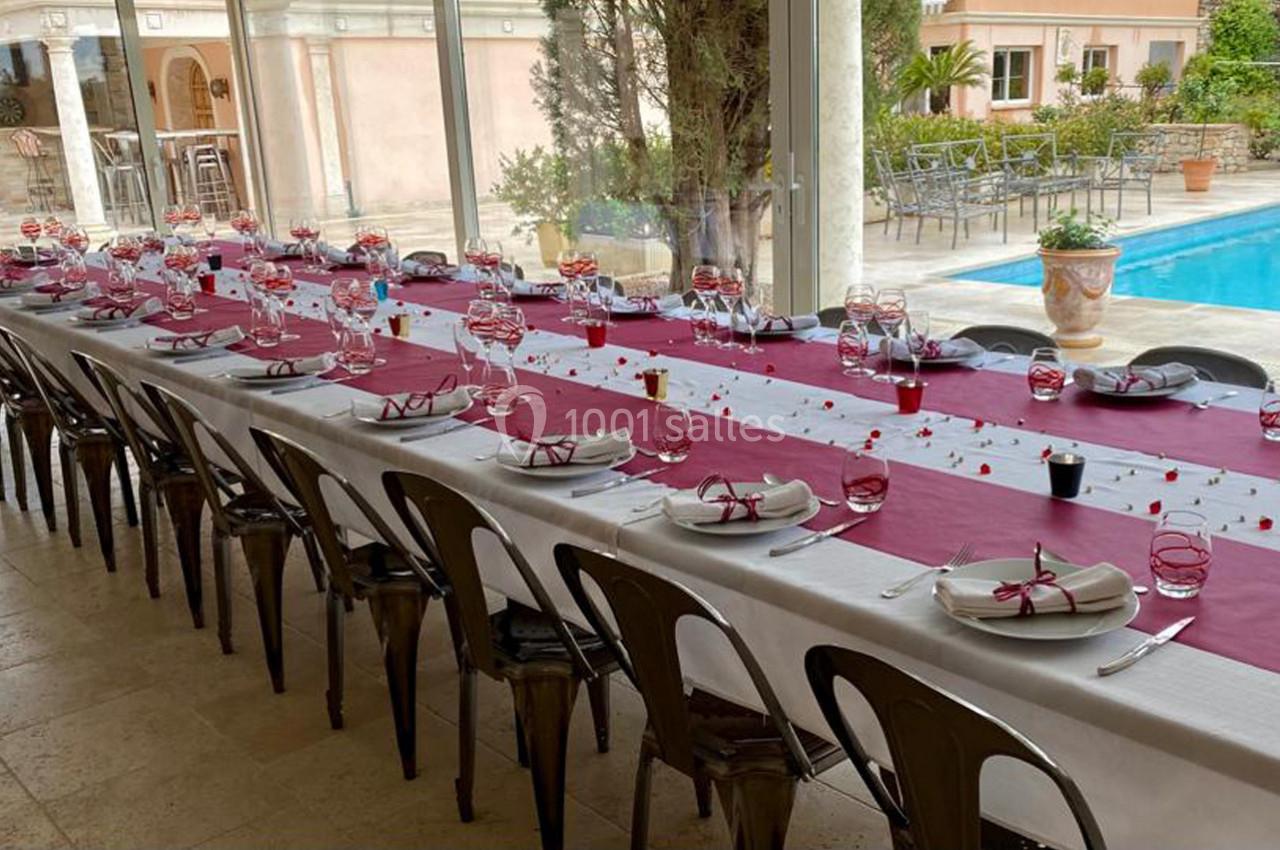 Table longue dressée pour un repas, nappes blanches et bordeaux, vaisselle élégante, près d'une piscine extérieure.