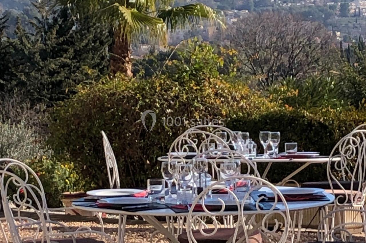 Tables en fer forgé dressées avec vaisselle et verres, situées en extérieur dans un jardin avec vue sur des collines.