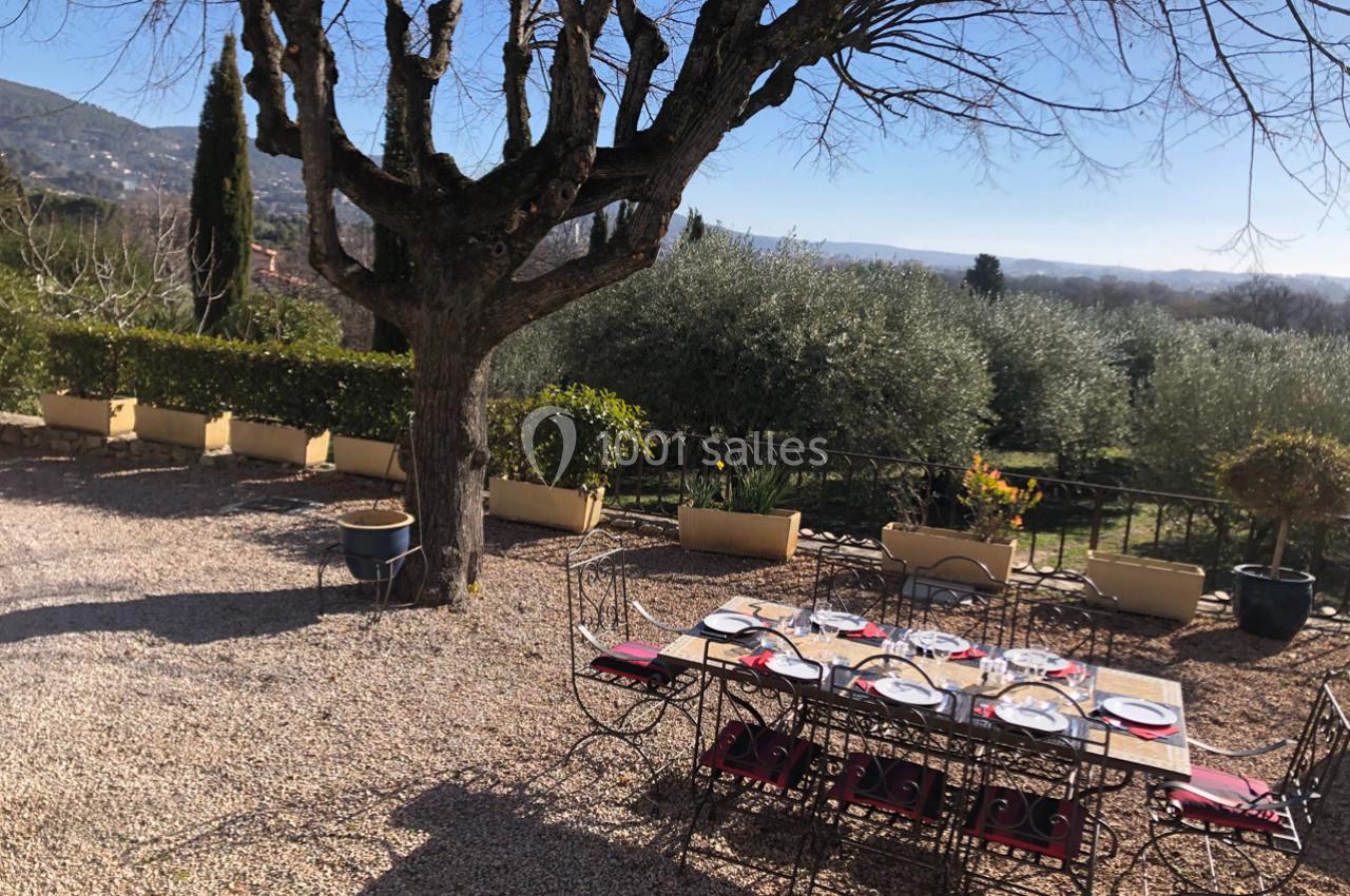 Table dressée en extérieur sous un arbre, entourée de gravier, avec vue sur un jardin et des oliviers.