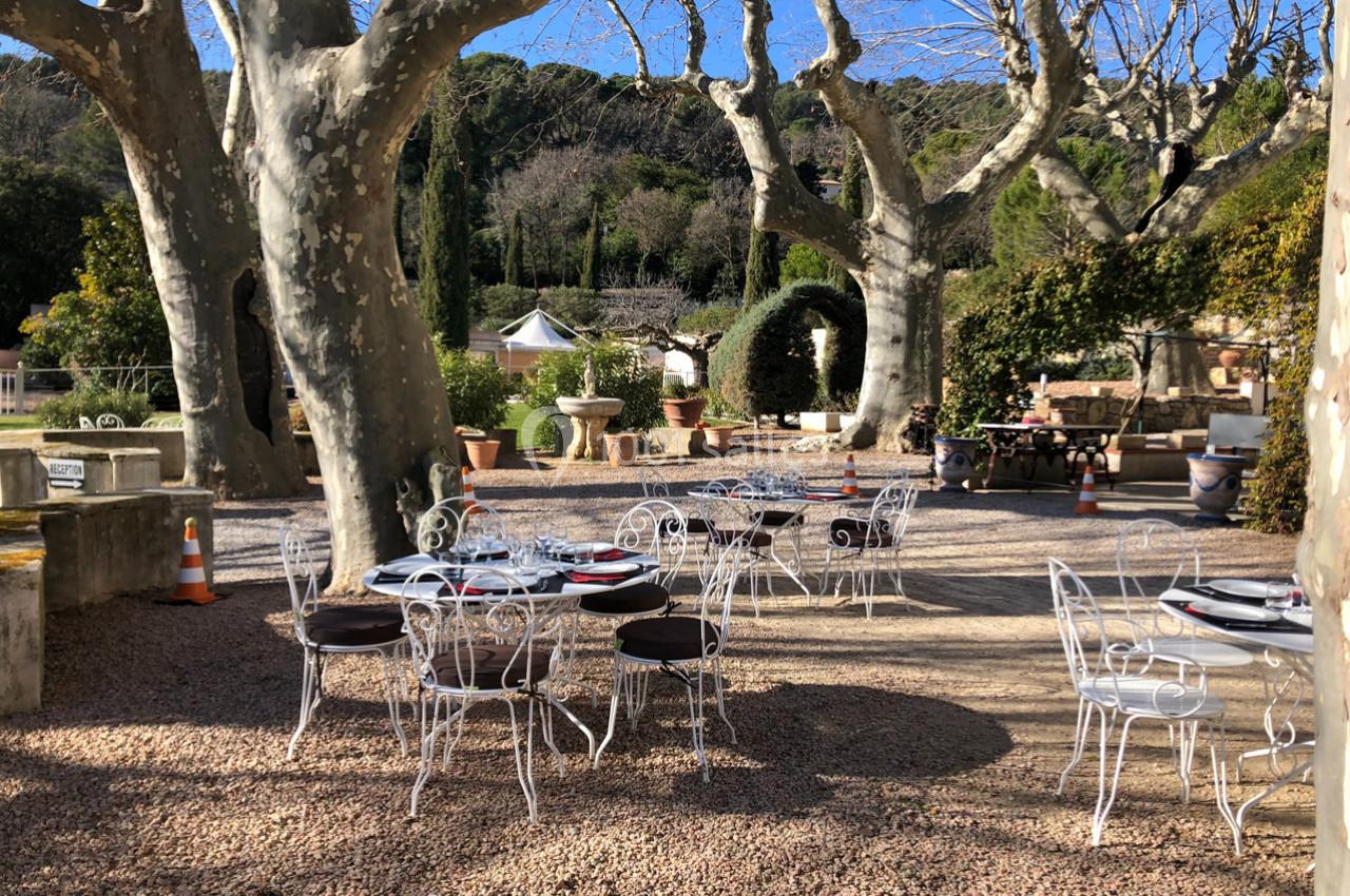 Tables et chaises en fer forgé disposées sur une terrasse en gravier, entourées d'arbres et de végétation.