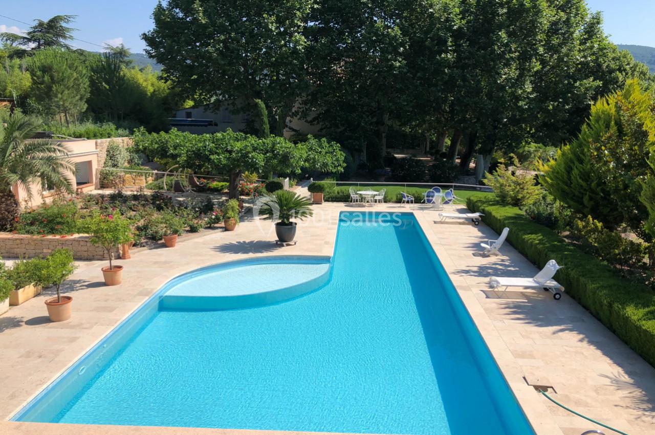 Piscine extérieure rectangulaire entourée de végétation, de chaises longues et de pots de fleurs sur une terrasse en pierre.