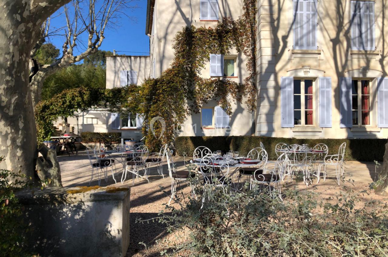 Terrasse ensoleillée avec tables et chaises en fer forgé devant une maison aux murs recouverts de végétation.