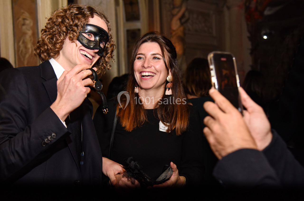 Un homme en costume portant un masque et une femme souriante posent pour une photo lors d'un événement festif.