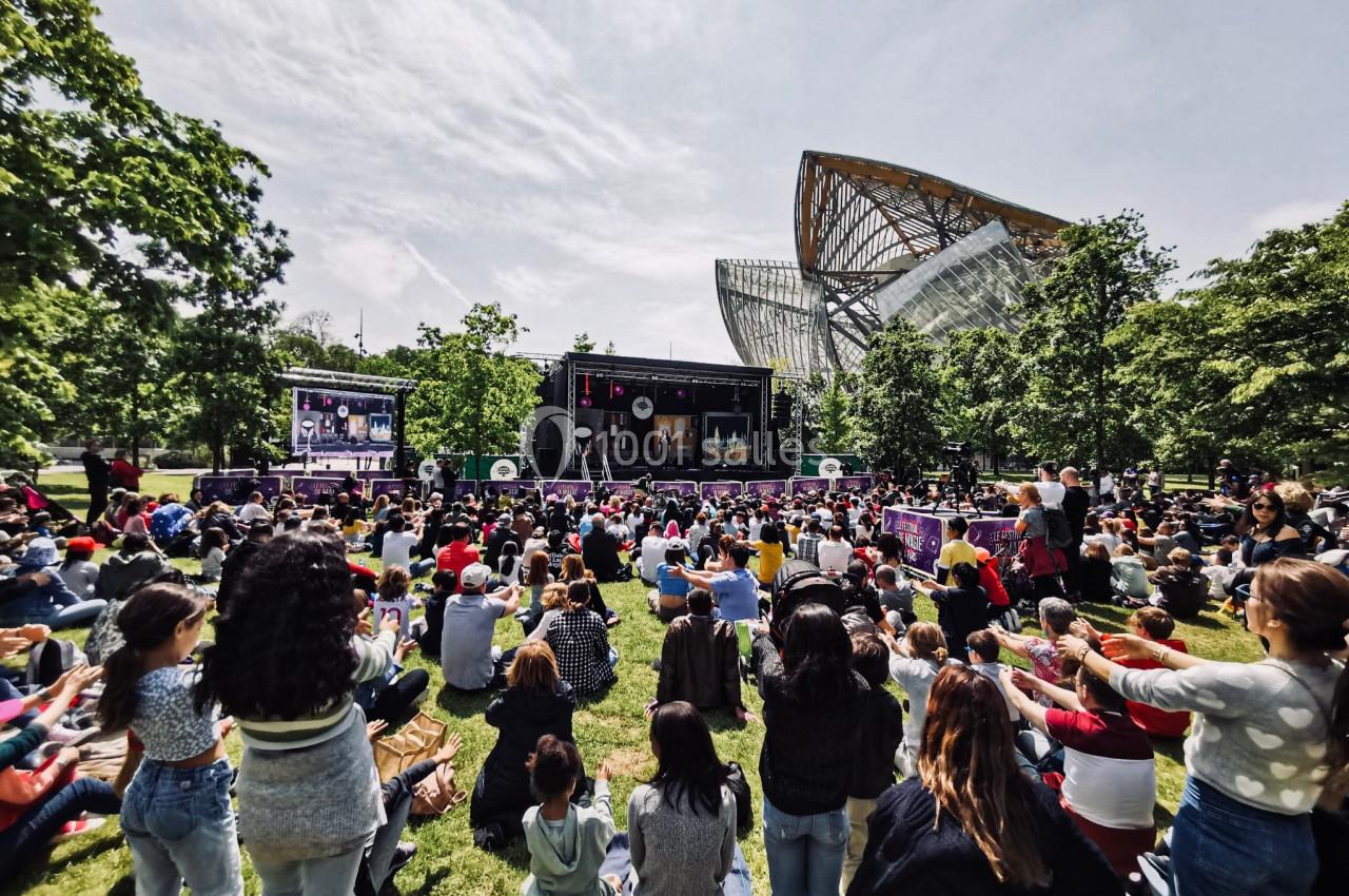 Foule assise sur une pelouse devant une scène en plein air, entourée d'arbres et d'une architecture moderne.
