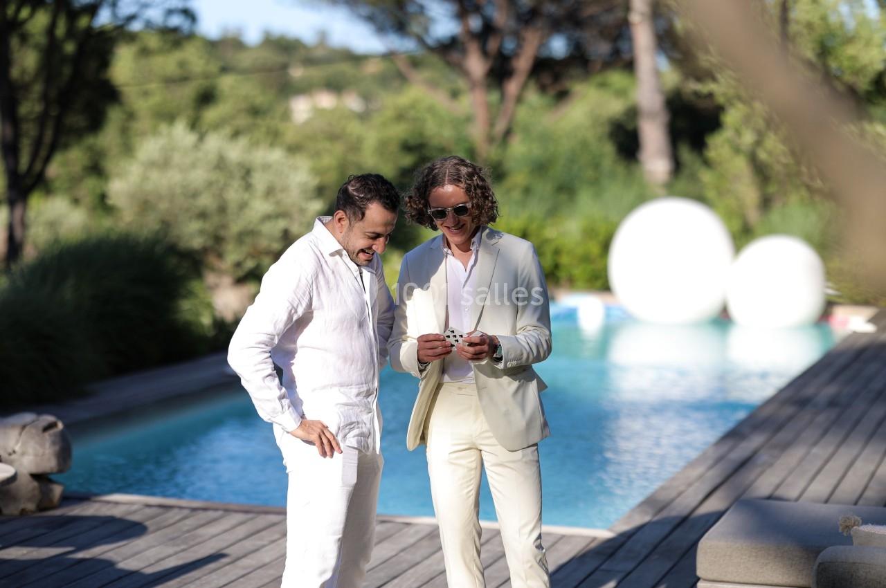 Deux hommes en tenue élégante discutent près d'une piscine entourée de végétation.