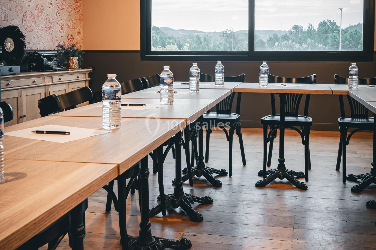 Salle de réunion avec tables en bois, chaises noires, bouteilles d'eau et carnets, éclairée par une grande fenêtre.