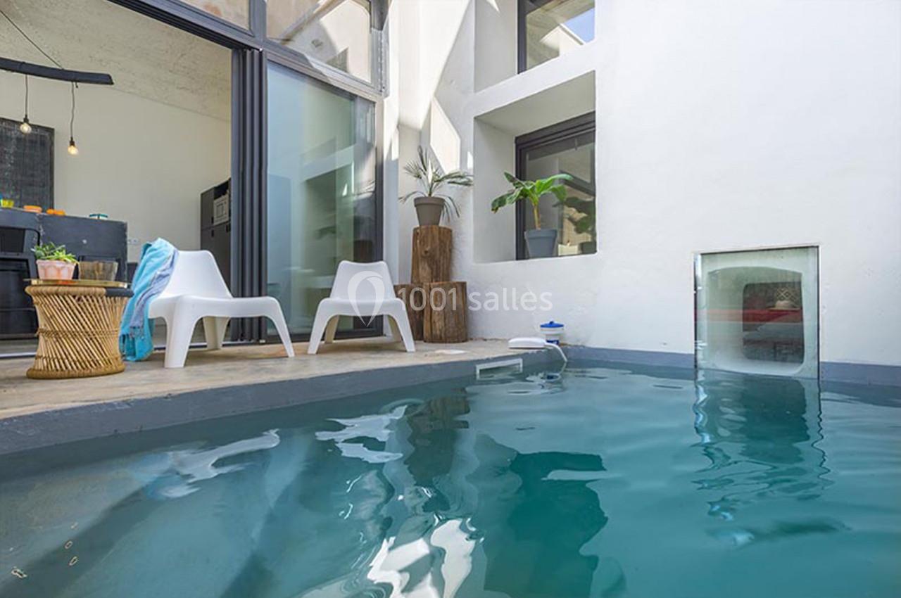 Piscine intérieure moderne avec chaises blanches, table en osier et plantes près de grandes baies vitrées.