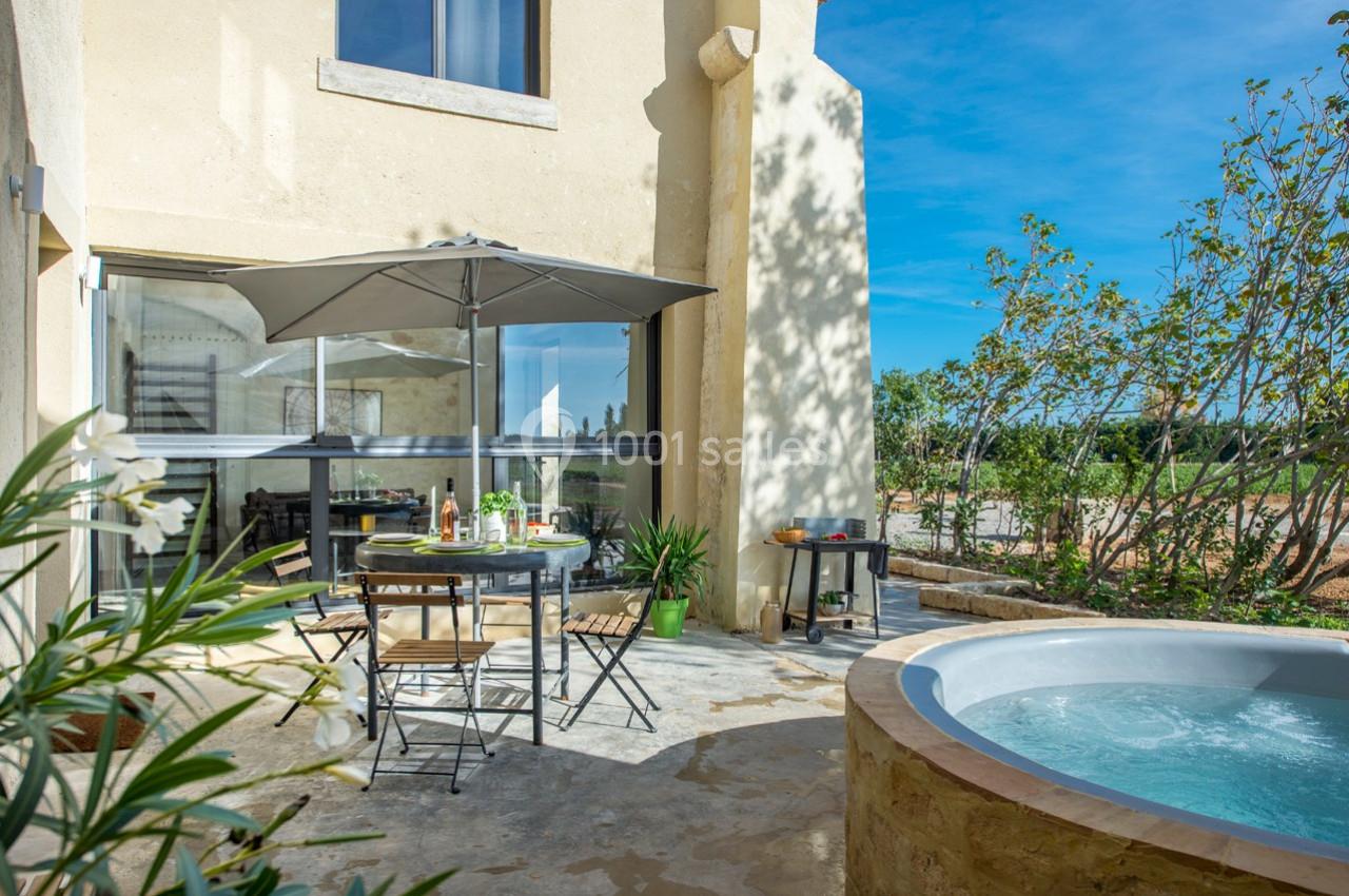 Terrasse ensoleillée avec table, chaises, parasol et jacuzzi, entourée de verdure et d'un bâtiment en pierre.