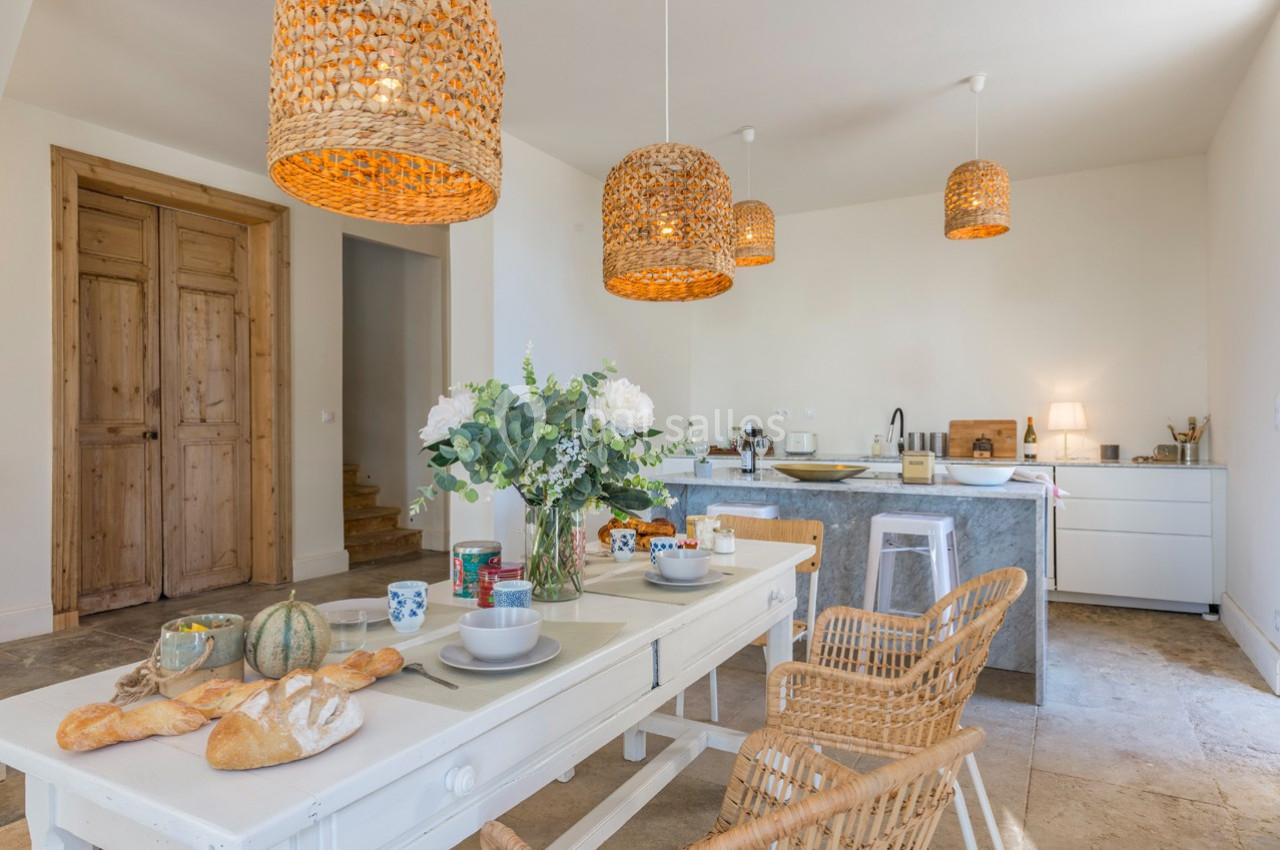 Cuisine lumineuse avec table dressée, chaises en rotin, suspensions en osier et plan de travail moderne en arrière-plan.