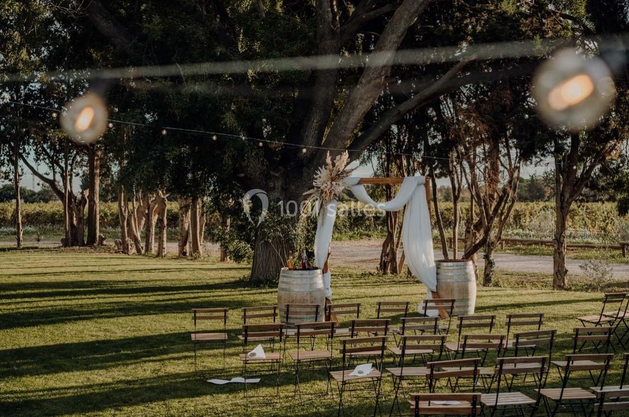 Chaises disposées en rangées devant une arche décorée de voilages et fleurs, installée dans un jardin sous des arbres.