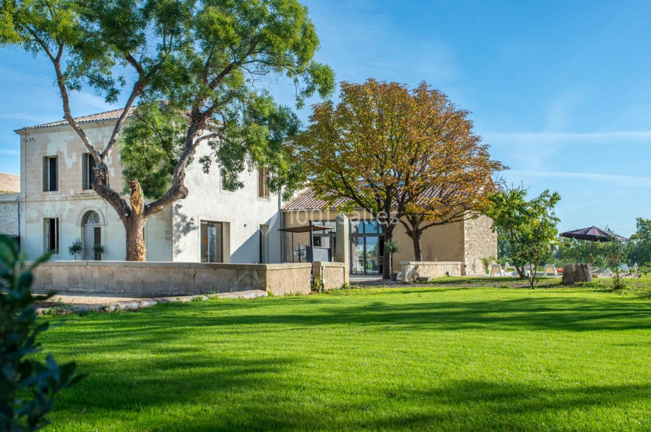 Maison en pierre entourée d'un jardin verdoyant, avec des arbres et un ciel bleu en arrière-plan.