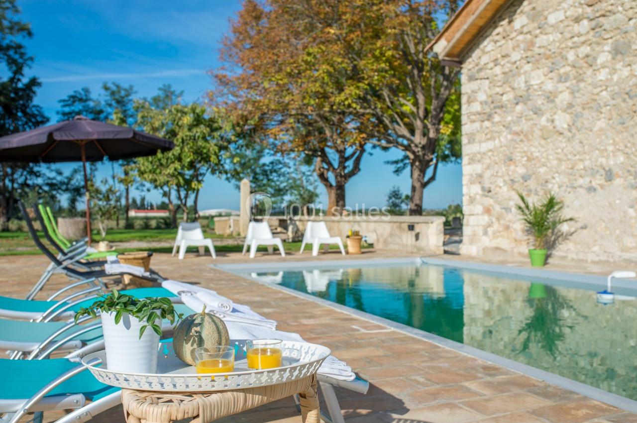 Terrasse avec piscine, transats, parasol et table avec jus d’orange, entourée de verdure et d’un bâtiment en pierre.