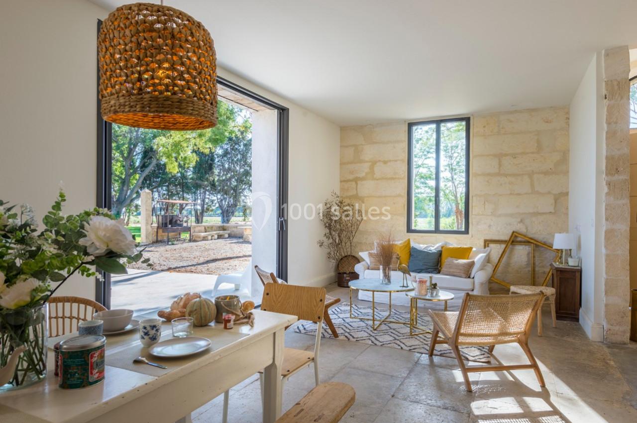 Salon lumineux avec murs en pierre, mobilier en bois clair, grandes baies vitrées et vue sur un jardin arboré.