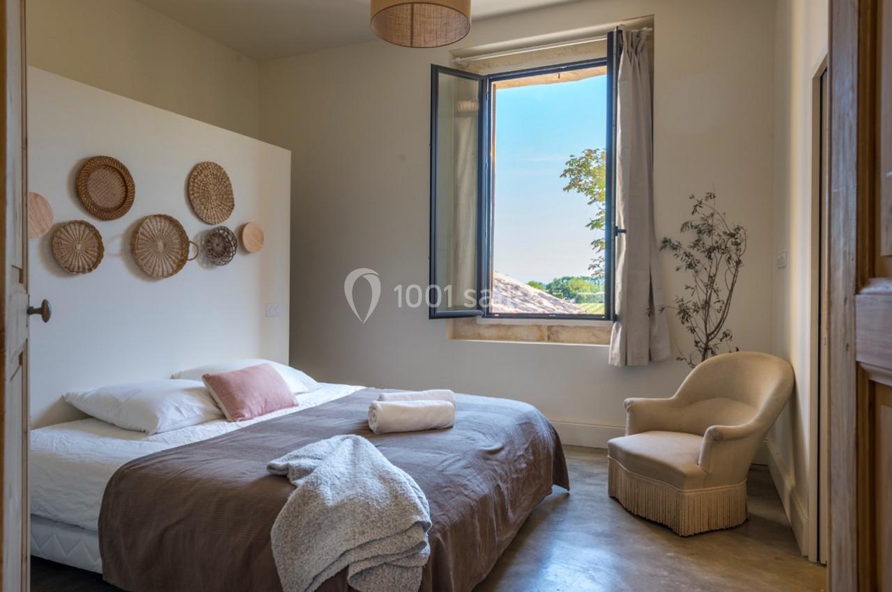 Chambre lumineuse avec lit double, fauteuil, décoration murale en osier et fenêtre ouverte sur un paysage verdoyant.