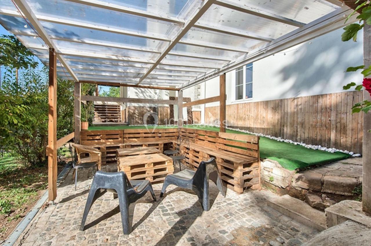 Terrasse extérieure avec mobilier en palettes, chaises en plastique et toit transparent, entourée de verdure.