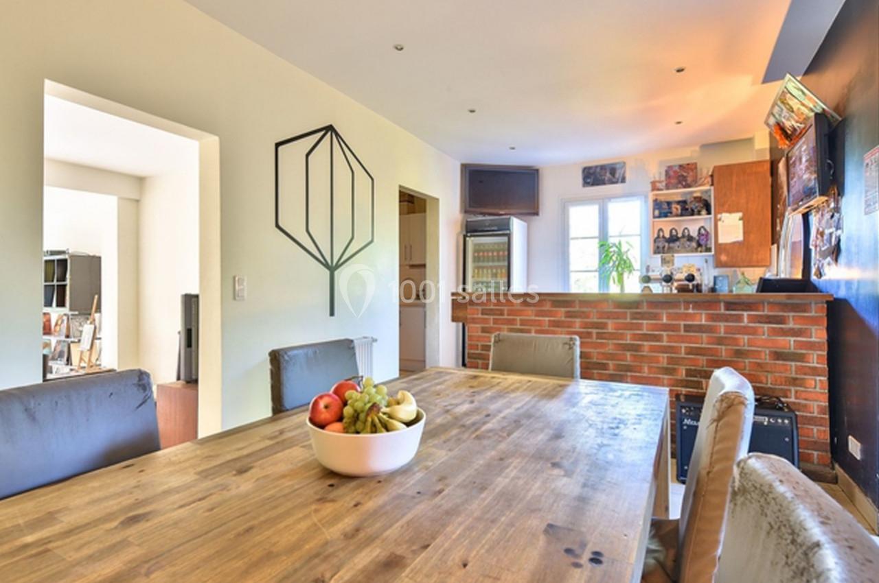 Salle à manger lumineuse avec table en bois, bol de fruits, chaises rembourrées et cuisine ouverte en briques au fond.